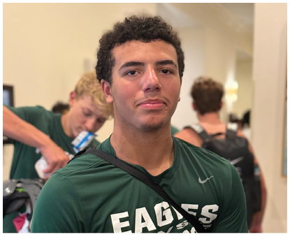 A person wearing a green shirt with the word Eagles stands indoors with two other people in the background, one pouring a drink. Description generated by AI
