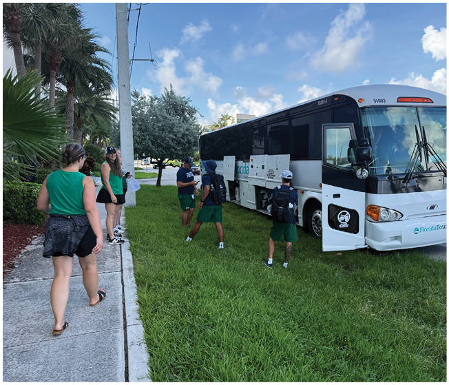 Several people wearing green shirts and shorts are standing and walking near a white and black tour bus parked on grass beside a sidewalk lined with palm trees under a partly cloudy sky. Description generated by AI