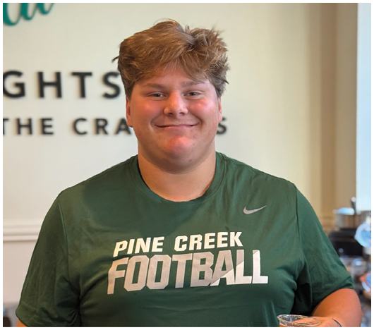 A person with short hair wearing a green Pine Creek Football t-shirt is smiling and holding a cup of iced tea in a kitchen setting. Description generated by AI