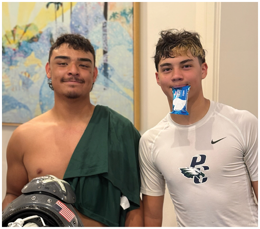 Two people stand indoors with one wearing a green draped cloth and the other in a white athletic shirt holding a blue and white snack in their mouth. Description generated by AI