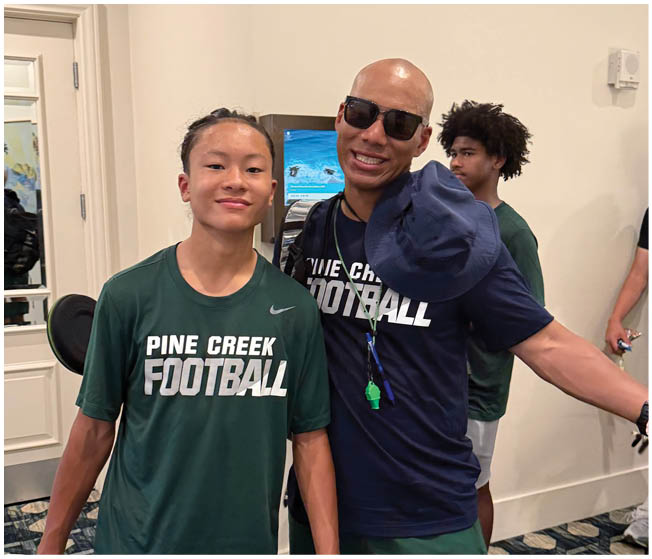 Two adults wearing Pine Creek Football shirts pose together indoors while two other people stand in the background. Description generated by AI