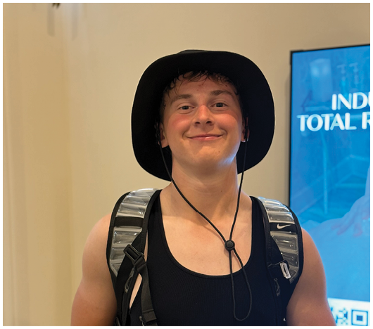 A person wearing a black tank top, a wide-brimmed hat, and a backpack is smiling in front of a blue sign. Description generated by AI