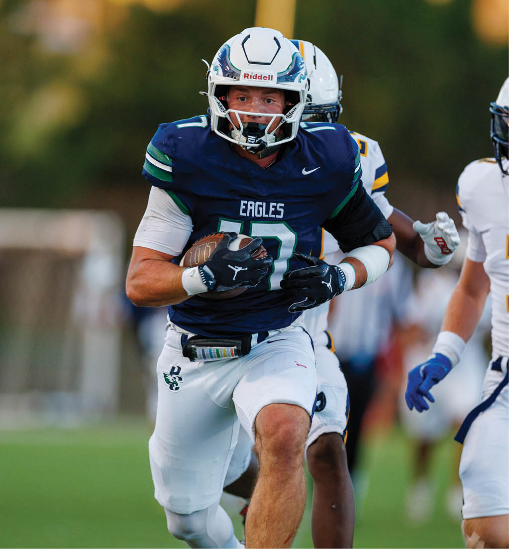 A person wearing a blue football uniform with the word Eagles is running on a field while holding a football, with other players visible in the background. Description generated by AI