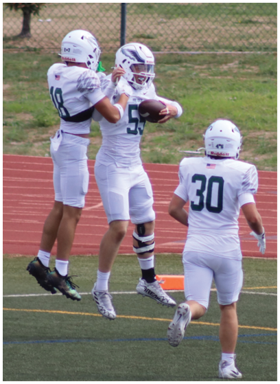 Three people in white football uniforms and helmets are on a field, with one person holding a football and another adjusting their helmet. Description generated by AI