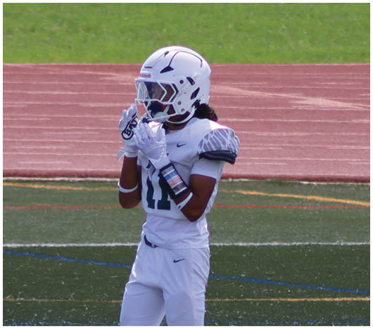 A person wearing a white football uniform and helmet stands on a field with a running track in the background. Description generated by AI
