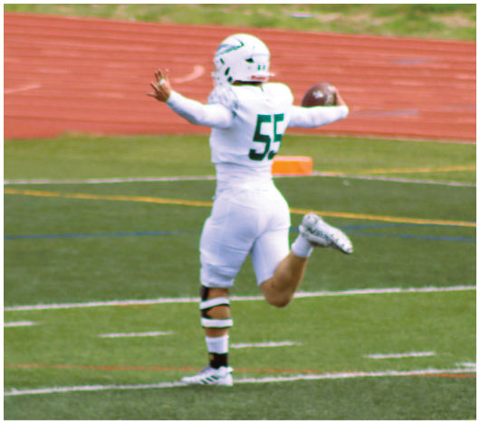 A person wearing a white football uniform with the number 55 is holding a football and appears to be preparing to throw it on a sports field with a running track in the background. Description generated by AI