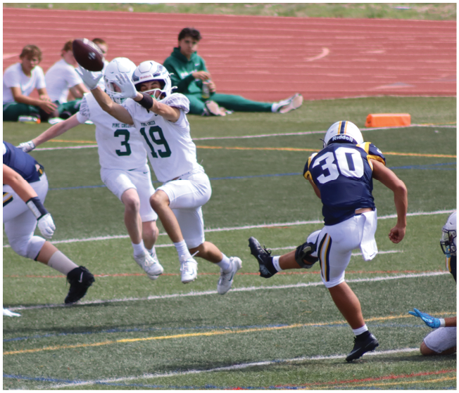 A person in a blue and white football uniform is kicking a ball held by another person kneeling on the ground while two people in white uniforms jump to block the kick on a sports field. Description generated by AI