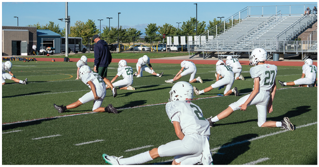 People in white football uniforms are stretching on a green field with a coach standing nearby and empty bleachers in the background. Description generated by AI