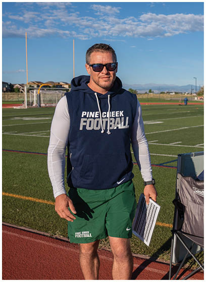 A person wearing a white football uniform and helmet is holding a football while standing on a grassy field near a running track. Description generated by AI