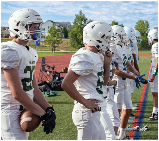 Several people in white football uniforms and helmets stand in a line on a grassy field near a running track, with one holding a football. Description generated by AI