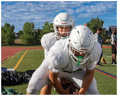 Two people in white football uniforms and helmets are engaged in a practice drill on a grassy field near a running track. Description generated by AI