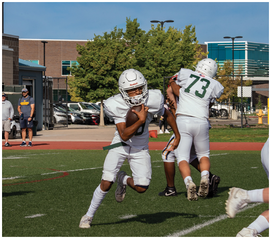 A person in white football gear is running with a football on a field while two people stand in the background near a track and parked cars. Description generated by AI
