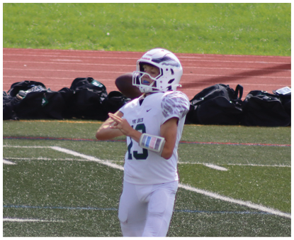 A person wearing a white football uniform and helmet is preparing to throw a football on a sports field with a running track and bags in the background. Description generated by AI