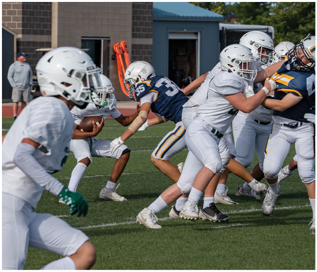 People wearing football uniforms are actively engaged in a play on a grassy field with one person holding the ball and others reaching or running nearby. Description generated by AI