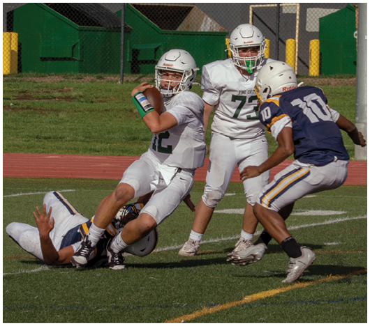 Two people in football uniforms are on the ground during a game on a grassy field near a running track. Description generated by AI
