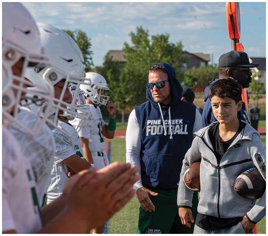 A group of people wearing white football helmets and uniforms stand in a line on a grassy field, with one person clapping in the foreground. Description generated by AI