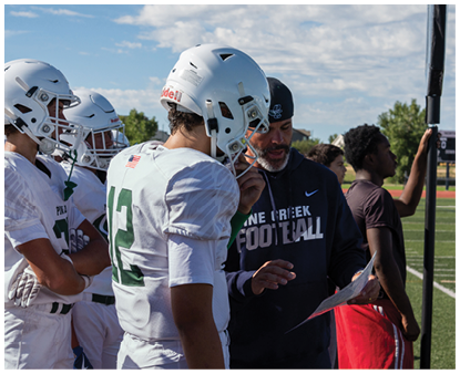 People in football uniforms and helmets are gathered around a person in a dark hoodie who appears to be giving instructions or discussing a play. Description generated by AI