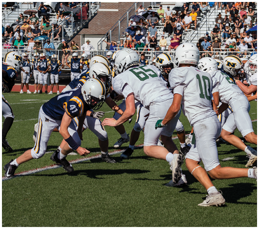 Two teams of people in football uniforms are engaged in a play on a grassy field with one player in a navy blue uniform crouching and reaching toward the ground while players in white uniforms approach. Description generated by AI