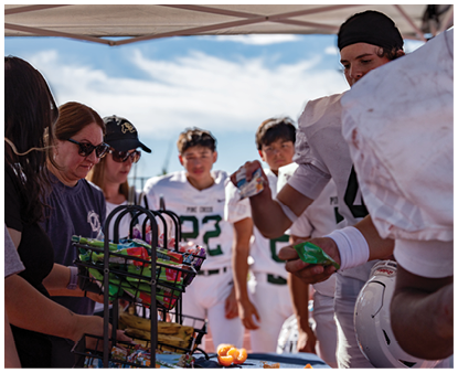 People in sports uniforms and casual clothing are gathered around a table with baskets of snacks outdoors on a sunny day. Description generated by AI