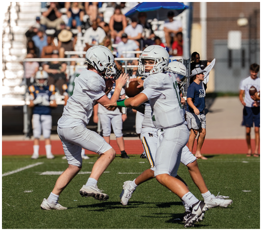 People wearing white sports uniforms are playing on a grassy field with a blurred background of other players and a track. Description generated by AI