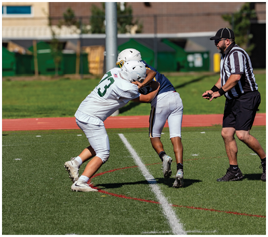 Two people wearing helmets and sports uniforms are engaged in a physical play on a grassy field marked with white and red lines. Description generated by AI