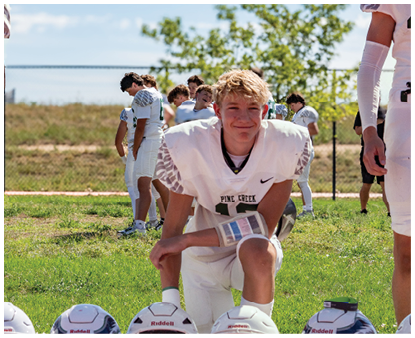 A person in a white football uniform kneels on grass with several helmets lined up in front while other people in similar uniforms stand and talk in the background near a fence and trees. Description generated by AI