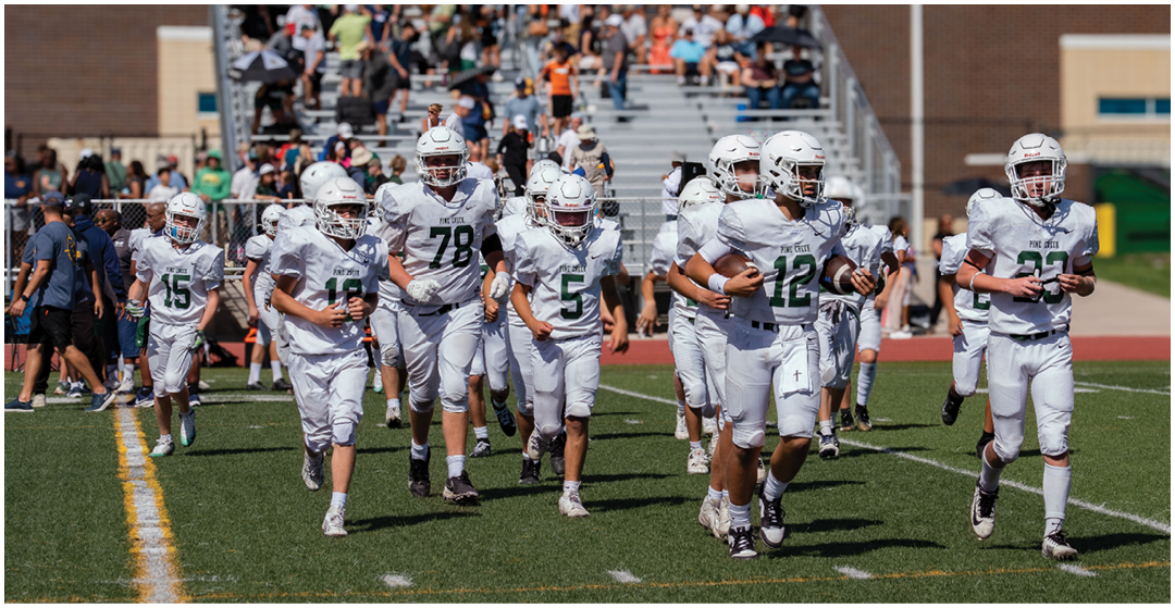A group of people wearing white football uniforms and helmets are running on a green field with spectators sitting on bleachers in the background. Description generated by AI