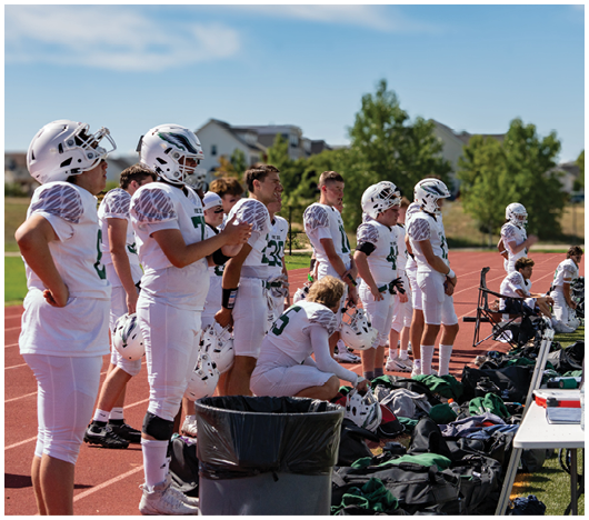 People in white football uniforms stand and sit on the sidelines of a sports field with helmets and gear scattered around. Description generated by AI