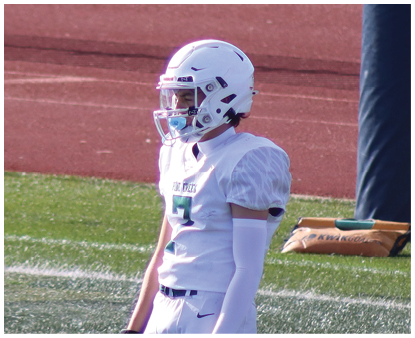A person wearing a white football uniform and helmet stands on a field with green turf and a red running track in the background. Description generated by AI