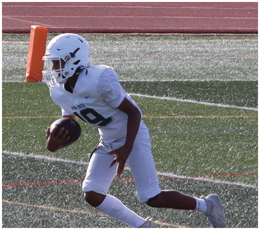 A person in a white football uniform and helmet is holding a football and appears to be running on a field near an orange pylon. Description generated by AI
