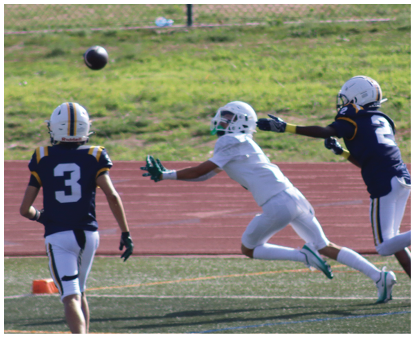 Two children in football uniforms are reaching for a football in midair on a field near a running track. Description generated by AI