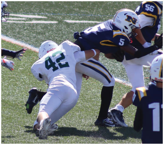 Two people in football uniforms are engaged in a tackle on a grassy field with others visible in the background. Description generated by AI
