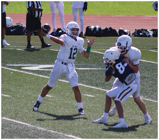 A person in a white football uniform is preparing to throw a football while another person in white tackles a person in a blue football uniform on a field. Description generated by AI