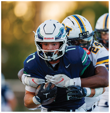 Two people in football uniforms are engaged in a tackle, with one holding the ball tightly while the other wraps arms around them. Description generated by AI