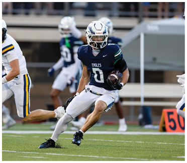 A person wearing a dark blue football jersey with the number zero and white pants is running on a football field while holding a football, with another player in white and yellow uniform nearby. Description generated by AI