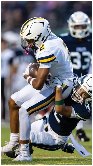 A person in a dark blue football uniform tackles another person in a white football uniform during a game on a grassy field. Description generated by AI