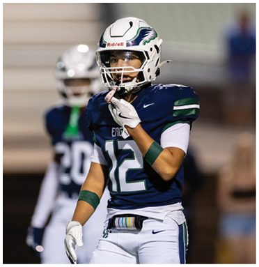 A person wearing a football uniform and helmet with the number 12 stands on a field with another player blurred in the background. Description generated by AI