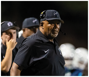 A person wearing a black cap and black sportswear stands on a football field holding a play sheet while other people in sports uniforms and headsets stand nearby. Description generated by AI
