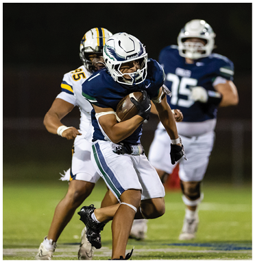 A person in a dark blue and white football uniform is running with the ball while being pursued by two other players in white uniforms on a football field at night. Description generated by AI