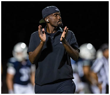 A person wearing a dark polo shirt and backward cap is clapping with a focused expression while standing on a sports field with blurred players in the background. Description generated by AI