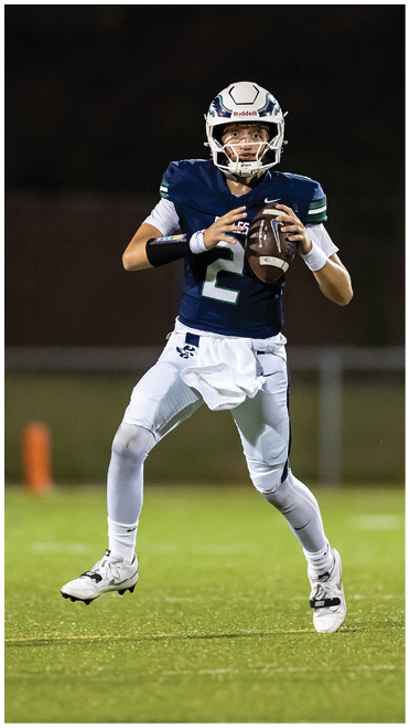 A person wearing a football helmet and navy blue jersey is holding a football in a ready-to-throw position against a dark background. Description generated by AI