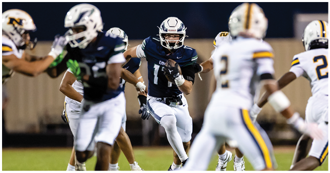 A person in a dark blue football uniform is running with the ball while several people in white and yellow uniforms attempt to block or tackle on a football field. Description generated by AI
