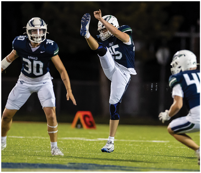 Two people in football uniforms are on a field with one person kicking a ball and the other person standing ready. Description generated by AI