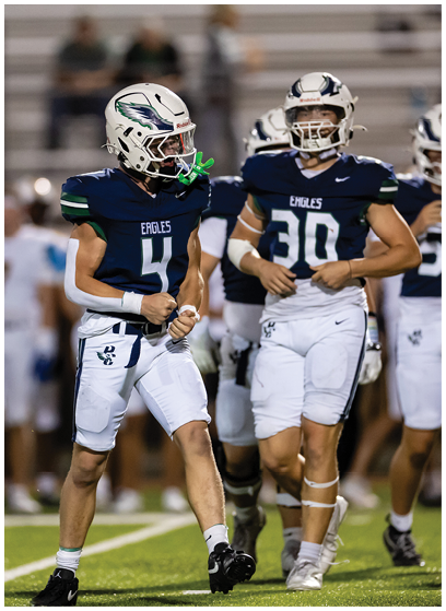 Two people in football uniforms with helmets stand on a field, one clenching fists in a celebratory pose while the other stands nearby. Description generated by AI