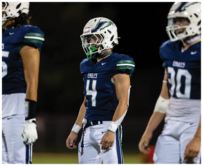 Three people wearing football uniforms and helmets stand on a field, with two facing forward and one partially visible on the right. Description generated by AI