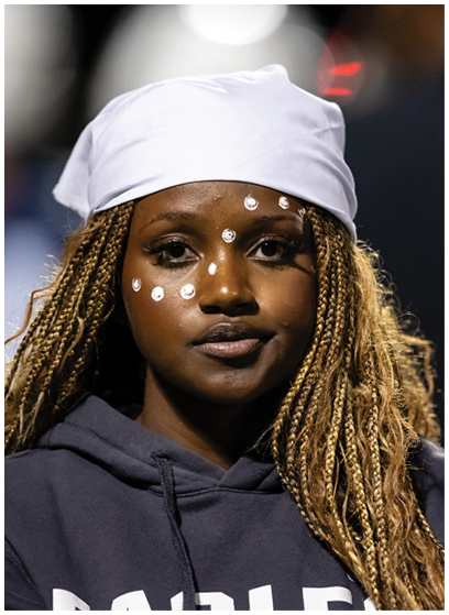 Person with long braided hair wearing a white headscarf and dark hoodie with white face paint dots on their face. Description generated by AI