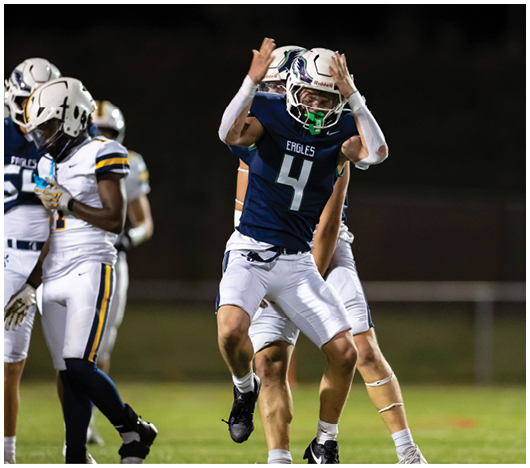 A person wearing a blue football jersey with the number 4 and the word Eagles is raising their arms while other players in white uniforms are nearby on a football field at night. Description generated by AI
