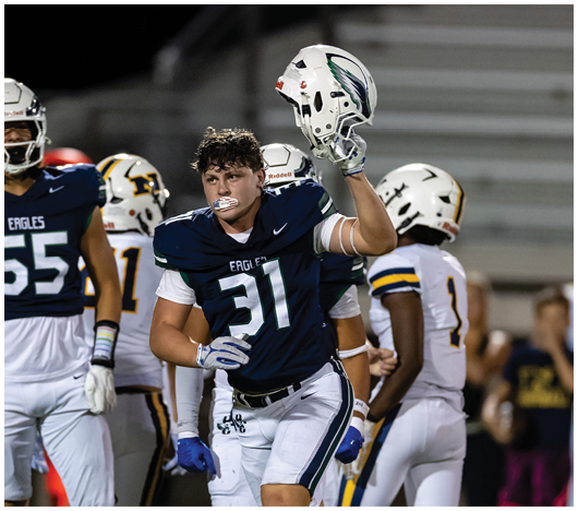 A person wearing a dark blue football uniform with the number 31 is holding up a white helmet while standing on a football field with other players in the background. Description generated by AI