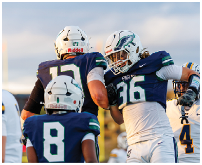 Three people wearing football helmets and navy blue and white uniforms are engaged in a celebratory gesture on a field during a game. Description generated by AI