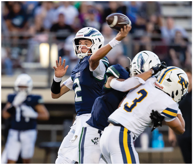 A person in a blue and white football uniform is throwing a football while another person in a white and yellow uniform attempts to block or tackle them during a game. Description generated by AI